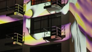 The building facade is lit by colourful cartoonish liquid colours. The projection covers a balcony in hues of purple, yellow, and white.
