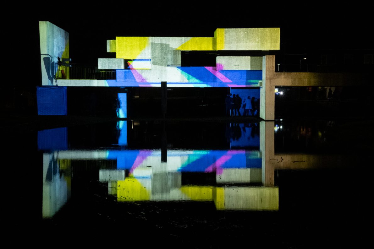 A concrete structure over water displays vibrant multi-coloured lights under a cloudy night sky with a bright moon. The building is mainly yellow with thick stripes of blue and thin pink stripes. The scene reflects on the calm water, creating a serene atmosphere.