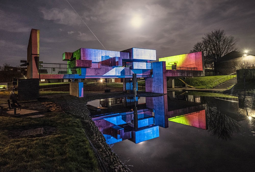 A concrete structure over water displays vibrant multi-coloured lights under a cloudy night sky with a bright moon. The building is covered in thick bands of red, yellow and purple lights - the whole structure is underlit with blue. The scene reflects on the calm water, creating a serene atmosphere.