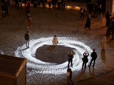 Light installation Hidden Voices: PROCESSIONS in the City at night in Aldgate Square. A spirograph pattern of white light projected on a Square at night