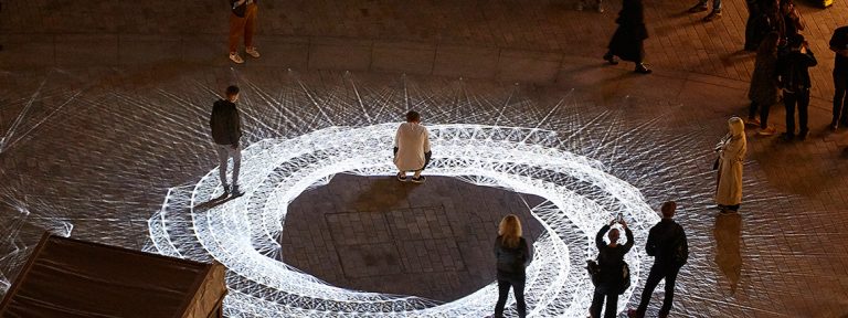 Light installation Hidden Voices: PROCESSIONS in the City at night in Aldgate Square. A spirograph pattern of white light projected on a Square at night
