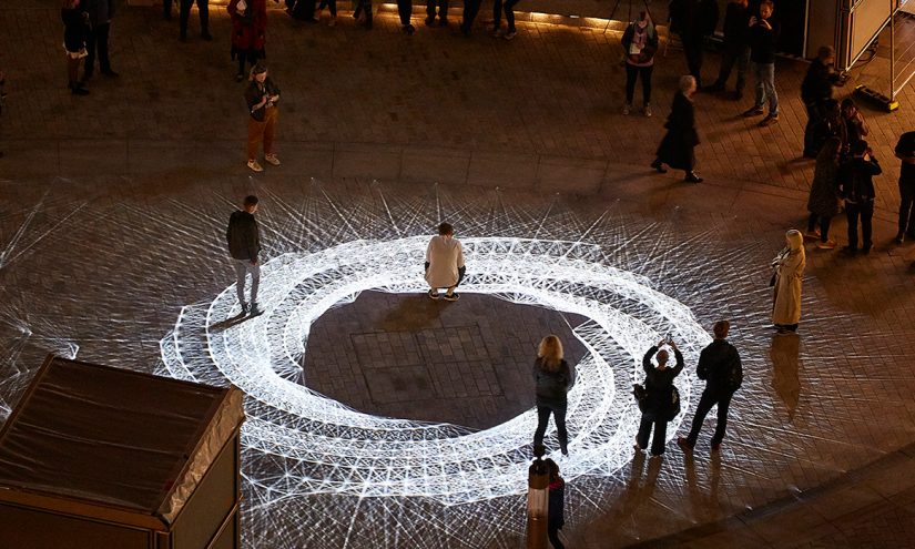 Light installation Hidden Voices: PROCESSIONS in the City at night in Aldgate Square. A spirograph pattern of white light projected on a Square at night
