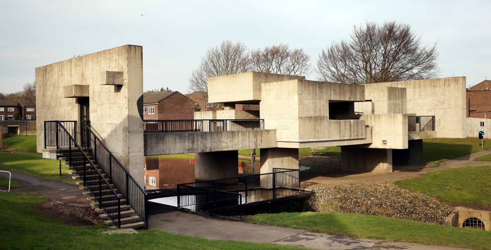 Brutalist concrete pedestrian bridges with angular structures cross a grassy area. Homes and bare trees are visible under an overcast sky.