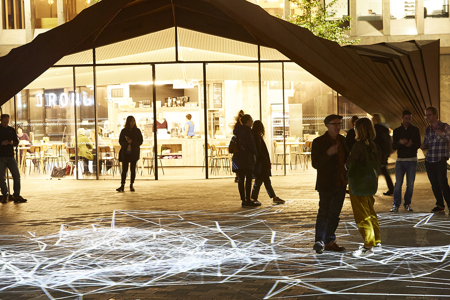 Contemporary outdoor pavilion with angular black geometric architecture illuminated at night in a busy urban plaza. Several people stand and socialise outside on the paved plaza, where abstract white light projections create intersecting line patterns across the ground.