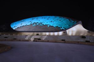 Autostadt pavilions in Germany. A domed structure is illuminated with blue, cartoonish, water-like projections at night. In the foreground, terraced seating creates a serene, futuristic ambiance.