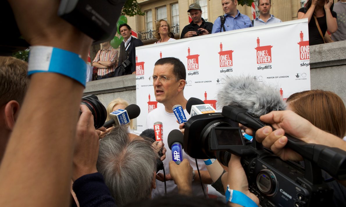 Antony Gormley surrounded by journalists with microphones. The backdrop reads One & Other in partnership with Sky Arts