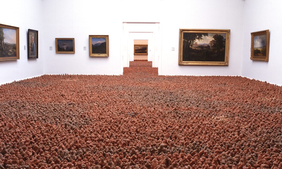 A white-walled gallery room filled with thousands of small brown clay figurines covering the floor. Framed paintings line the walls, creating a juxtaposition of classical and modern art. The scene evokes a sense of curiosity and wonder.