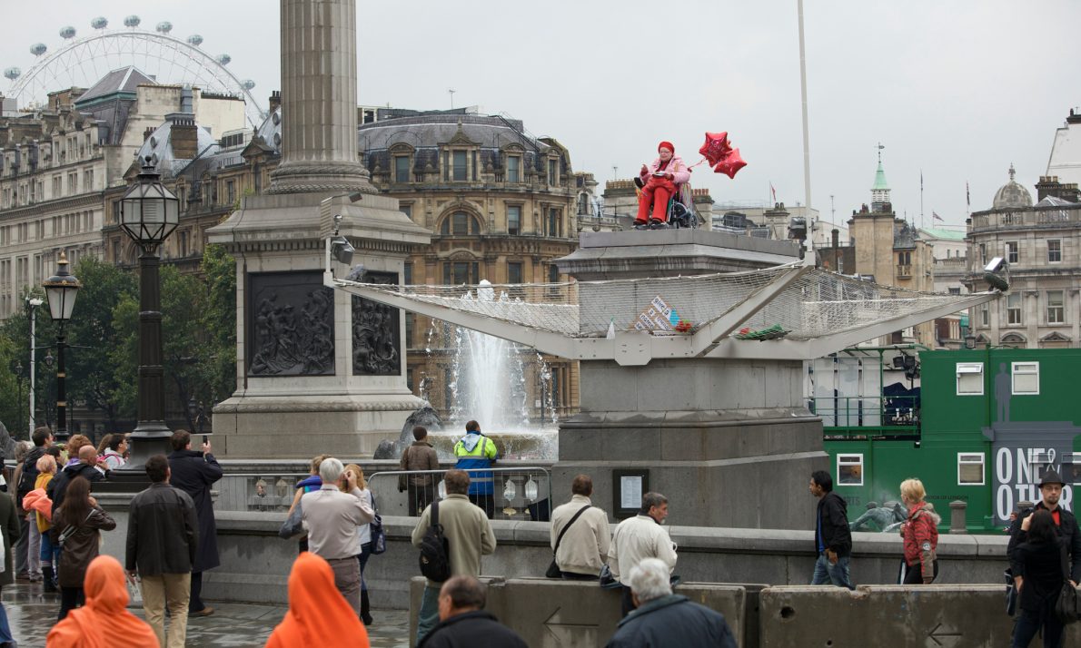 A stylish woman with a bald head is in a wheelchair is atop the Fourth Plinth in Trafalgar Square. She is wearing bright red and pink clothes and red star shaped balloons are tied to her chair. Her balloons read 