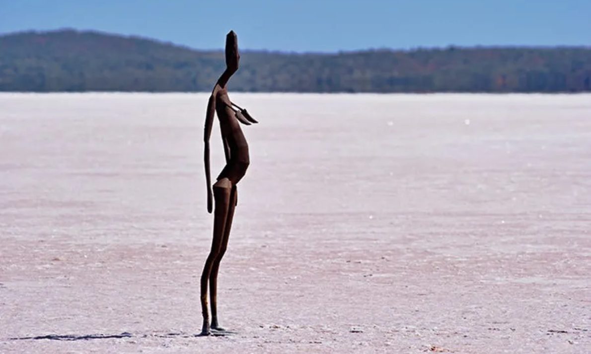 Sculpture of a slender, abstract human figure with a protruding stomach stands alone on a vast, dry salt flat under a clear blue sky. The sculpture is made from various brown metals. The work conveys feelings of solitude and serenity.
