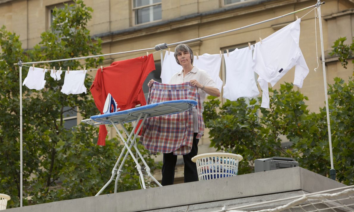 A person doing ironing on the Fourth Plinth with an ironing board and laundry basket. There is a washing line hung up behind them with various red and white clothes.