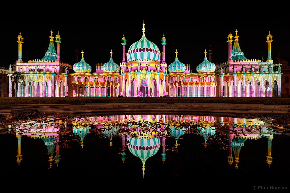 The image shows a night scene of Brighton’s Royal Pavilion Estate lit-up with colourful domes. The main body of the building is a mix of pink and yellow with deep orange patterns. The domes are various shades of blue and topped with yellow spires. The light is reflecting in a pool, creating a vibrant and festive atmosphere.