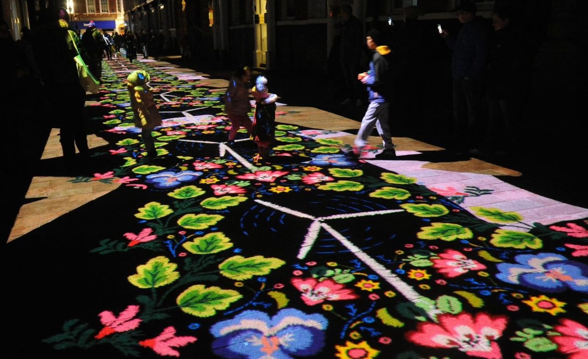 A dark street is illuminated by vibrant projections of pink and blue flowers and small bushy leaves. The graphics appear as if embroidered. There are children playing joyfully, creating a lively and whimsical atmosphere.