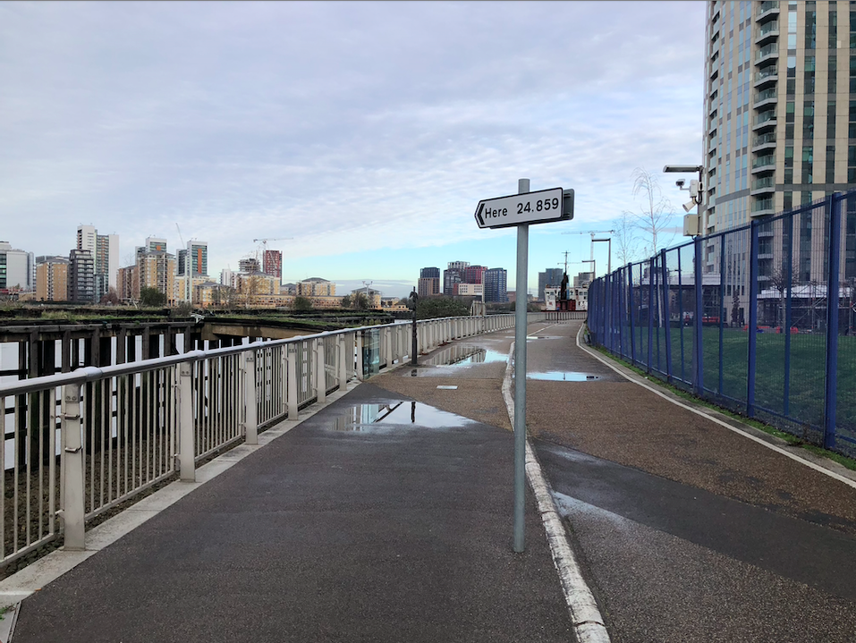 Here by Thomson & Craighead A walkway by the River Thames with a traffic sign planted in the middle, the sign is pointed in the direction of the river on it is written