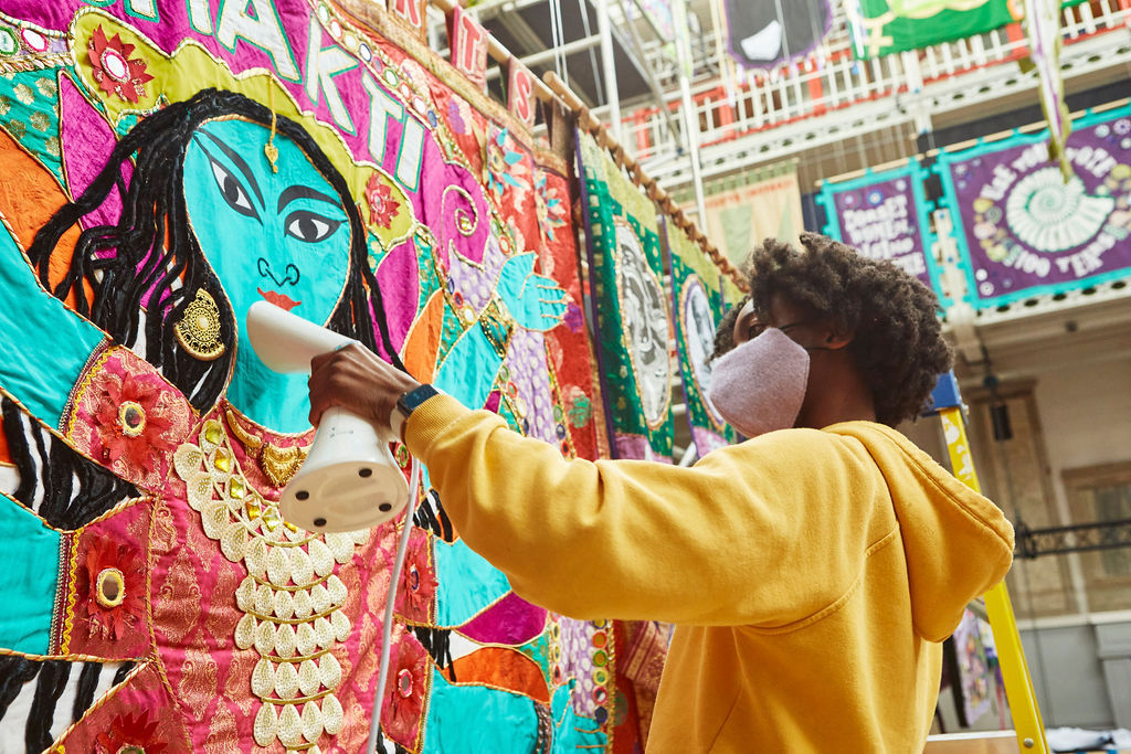 Communications Assistant, Khadija wearing a yellow hoodie and using a hand-held steamer to prepare a banner for the Women Making History exhibition.