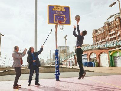 Man jumping in the air about to dunk a basketball into the net, on the right are two men one of which is holding up a microphone recording the sounds
