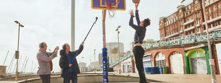 Man jumping in the air about to dunk a basketball into the net, on the right are two men one of which is holding up a microphone recording the sounds