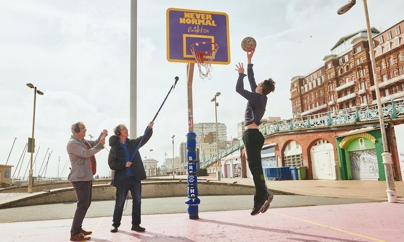 Man jumping in the air about to dunk a basketball into the net, on the right are two men one of which is holding up a microphone recording the sounds