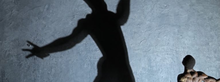 Man facing away and standing with his arms wide open - there is a giant shadow of him that is at the centre of the image