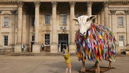 A giant sheep sculpture with a woolly coat made from hundreds of colourful fabric strips stands in a paved square in front of a grand sandstone building with tall Corinthian columns, part of Huddersfield railway station. A person in a yellow jacket and orange shorts reaches up towards the sheep's face, while two onlookers stand on the station steps in the background.