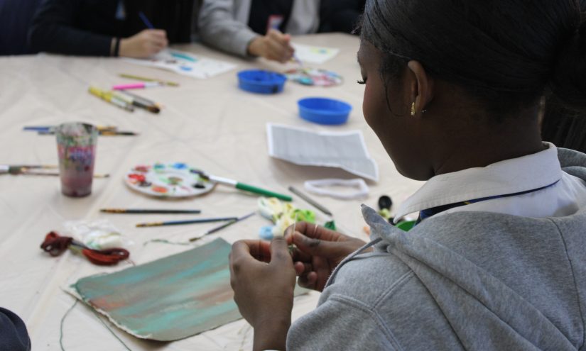 A young Black girl threads a needle whilst sitting at a table littered with art supplies.