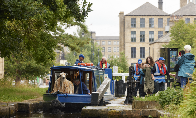 Pethera on the E-Shuttle on 14 July. HERD 2023, devised by Orlando Gough, produced by Artichoke, part of Kirklees Year of Music Photo by Matthew Andrews 4 The photograph features a canal boat with passengers and what seems to be a sheep-like figure on board. People, some wearing life jackets, are gathered around the canal and engaging with the scene, suggesting an interactive or performative element to the event.