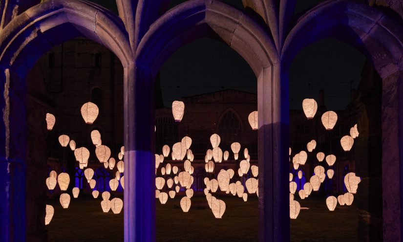Warmly-lit paper lanterns are seen in a courtyard at night through the cloisters of Durham Cathedral.