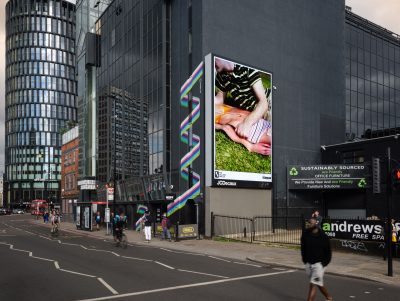 A city street with a large digital billboard displaying artwork of two people lying on grass.