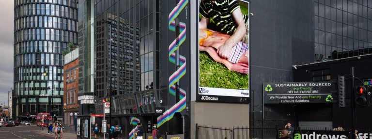 A city street with a large digital billboard displaying artwork of two people lying on grass.
