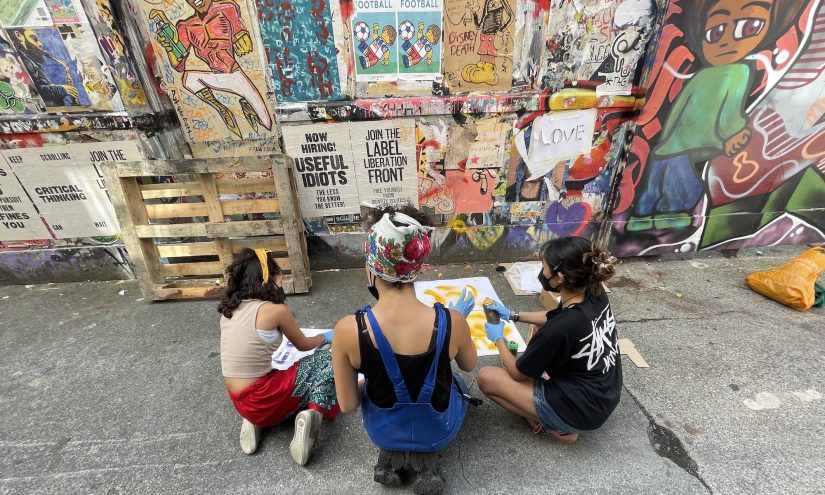 Three young individuals sit on the ground in front of a colorful graffiti-covered wall, engaging in a creative activity involving spray painting