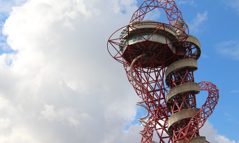 Red steel observation tower with a spiral slide against a partly cloudy sky, conveying a sense of modern architecture and adventure.