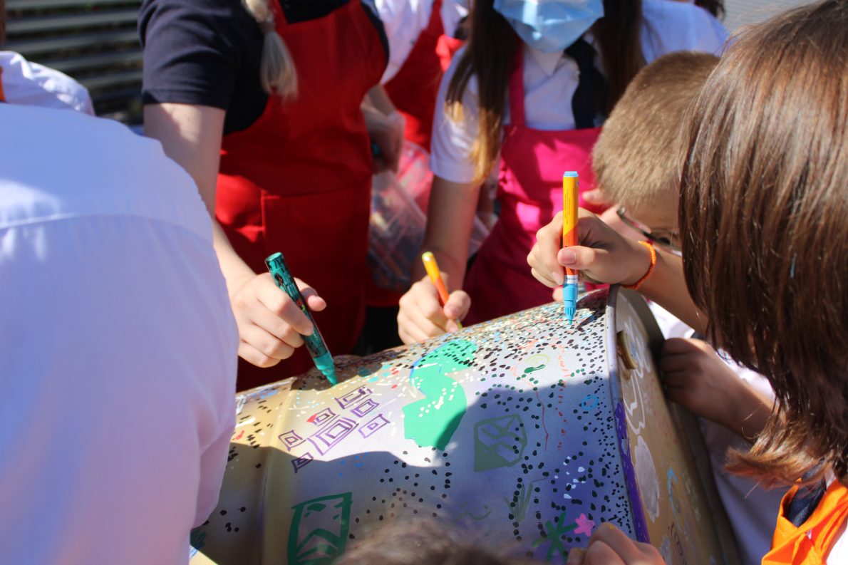 Children in aprons enthusiastically decorate a large metal sculpture with spray paints and colourful markers.