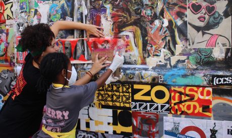 Tasnim Mahdy and a young participant collaborate to create street art on a colourful, graffiti-covered wall. One wears a mask as they spray the stencil with red spray paint. The scene is lively and expressive.
