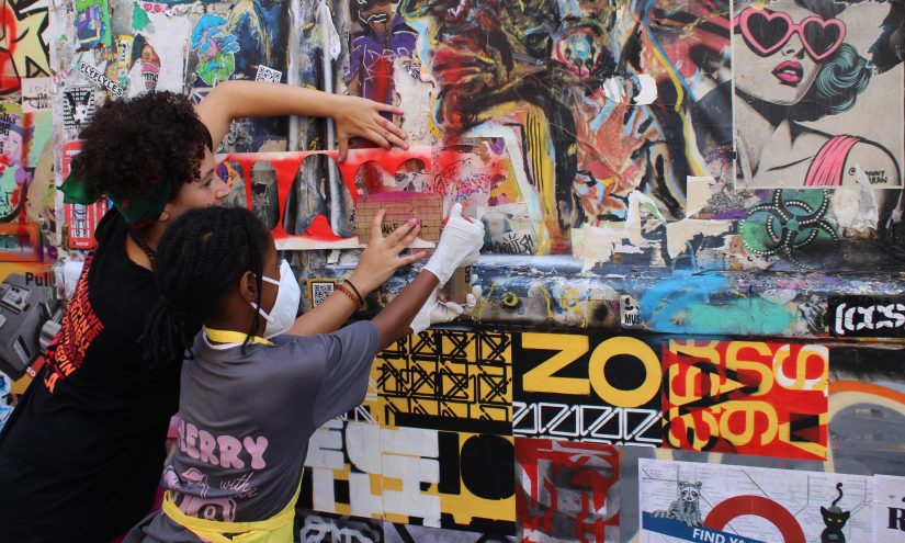 Tasnim Mahdy and a young participant collaborate to create street art on a colourful, graffiti-covered wall. One wears a mask as they spray the stencil with red spray paint. The scene is lively and expressive.