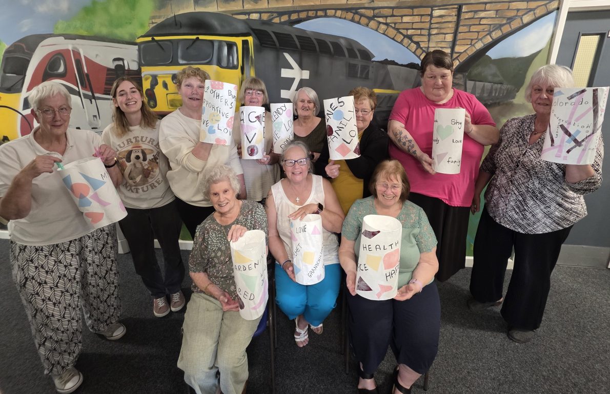 A group of smiling older women hold decorated paper lanterns with positive messages. They stand in front of a mural depicting trains and a bridge. The atmosphere is joyful