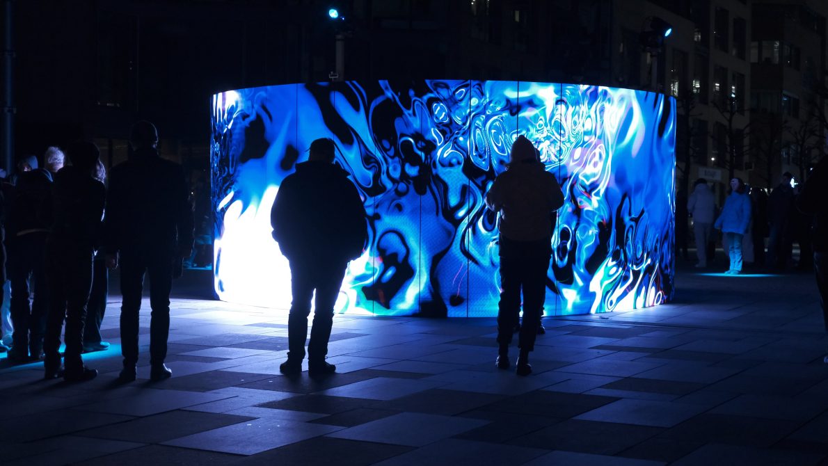 A crowd of people stands in a plaza at night, watching a large, curved LED screen. The screen is displaying a vibrant, abstract light show with swirling patterns in shades of electric blue and white. The figures in the foreground are silhouetted against the bright light of the screen, with the one in the center reaching out as if to touch it. In the background, there are other people, buildings, and a few small, glowing spotlights on the ground.