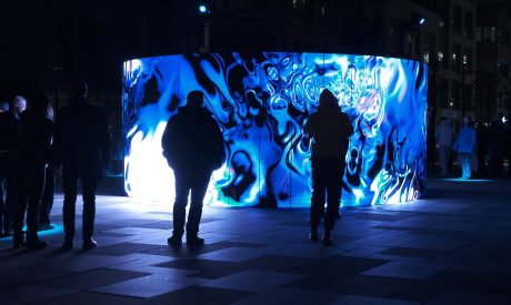 A crowd of people stands in a plaza at night, watching a large, curved LED screen. The screen is displaying a vibrant, abstract light show with swirling patterns in shades of electric blue and white. The figures in the foreground are silhouetted against the bright light of the screen, with the one in the center reaching out as if to touch it. In the background, there are other people, buildings, and a few small, glowing spotlights on the ground.