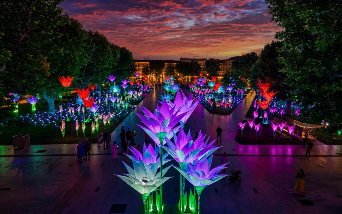 A vibrant night shot of a park filled with large, glowing floral light sculptures. In the foreground are several tall, blue and purple lotus-like flowers. A path extends into the distance, lined on both sides by trees and more glowing red, pink, and blue flowers. The sky is a dramatic mix of orange and purple as the sun sets. A few people are visible, walking along the path and enjoying the light installation.