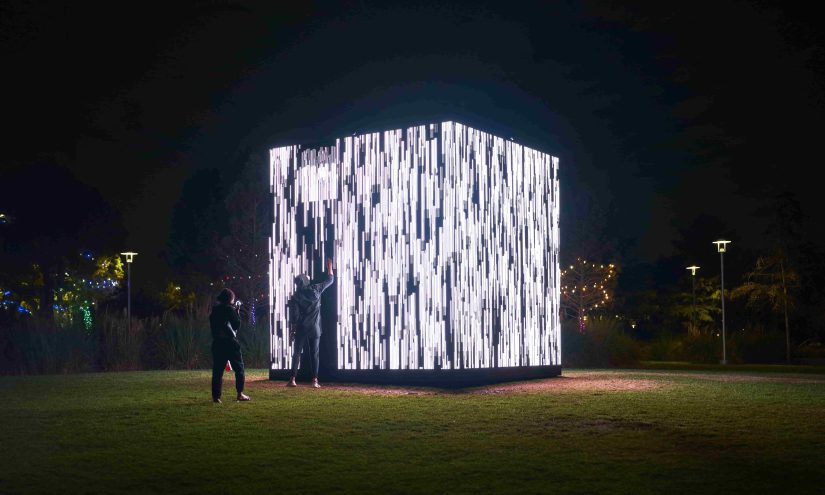A wide shot of two people interacting with a large, glowing cube on a grassy field at night. The cube is covered in vertical white lights that flicker and move like digital rain. The person on the left is holding a phone up to take a picture, and the person on the right is reaching out to touch the side of the cube. There are streetlights and trees in the background, dimly lit by the cube's glow and the distant lights of other installations.