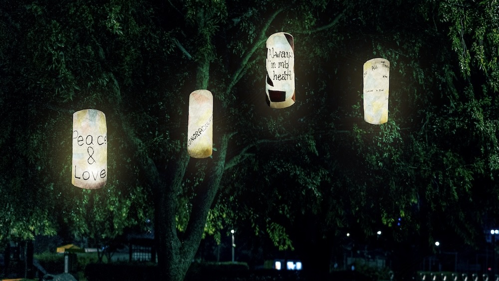 A nighttime photo of a park bench on a grassy field. Above the bench, hanging from the branches of a dark green tree, are four glowing cylindrical lanterns. Each lantern has different text or images, with 