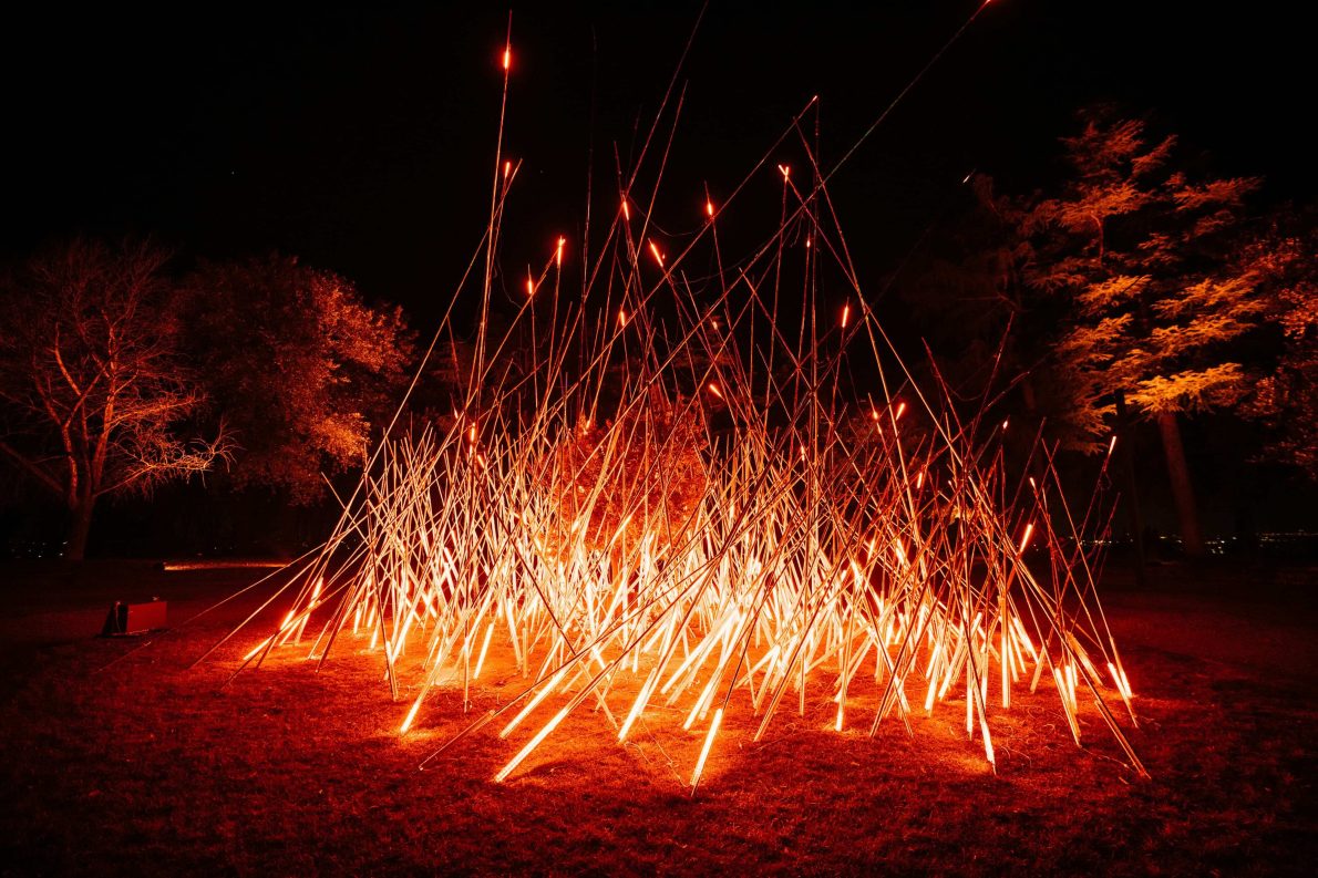 An outdoor light installation at night featuring a dense cluster of long, thin poles glowing bright orange-red. The illuminated poles crisscross upwards at sharp angles, creating the effect of fiery sparks shooting from the ground. Surrounding trees are faintly lit by the glow, contrasting against the dark night sky.
