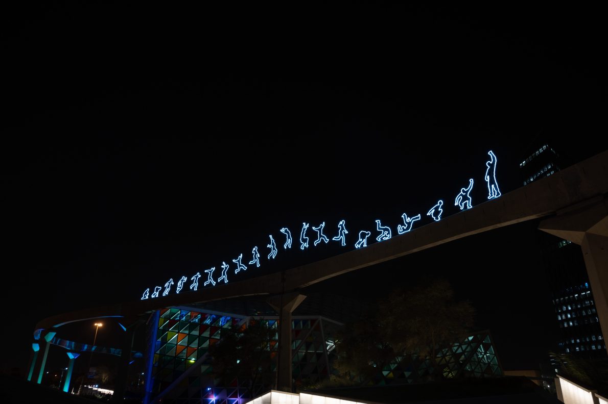 A long-exposure, night shot of a light installation. A row of illuminated, stick-figure-like silhouettes, in different poses, runs along a beam. The figures appear to be in a sequence of motion, from running to jumping. The figures glow with a bright blue light against a dark sky. Below the beam, a building with colourful geometric windows is visible.