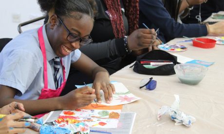 A girl wearing school uniform, glasses and smiling broadly, sits at a table and does art.