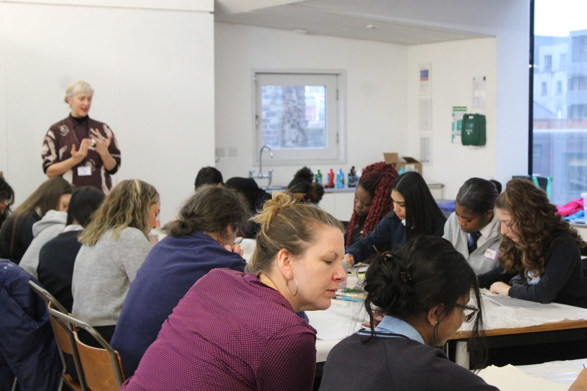 Students are sat at a large table working intently as an artist stands at the top, gesticulating.