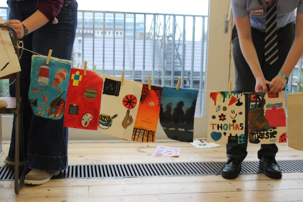 A selection of paintings made by young people are strung up and displayed in a studio.