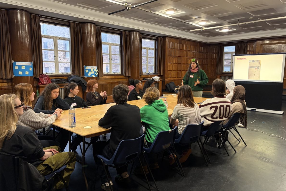 Students are sat at a large table listening intently to an artist, who stands at the top of the table wearing a green jumper and gesticulating.