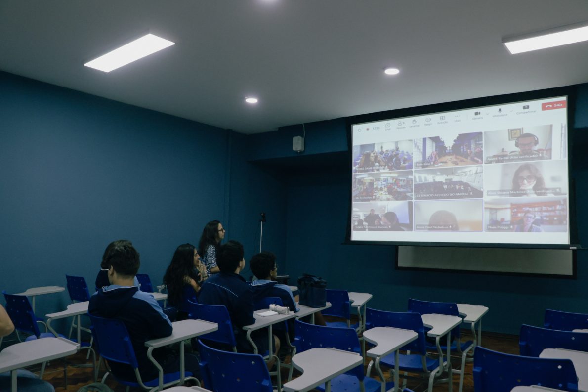 Students in a classroom look towards a projected screen of a Zoom call, where other schools and translators are presented on screen.