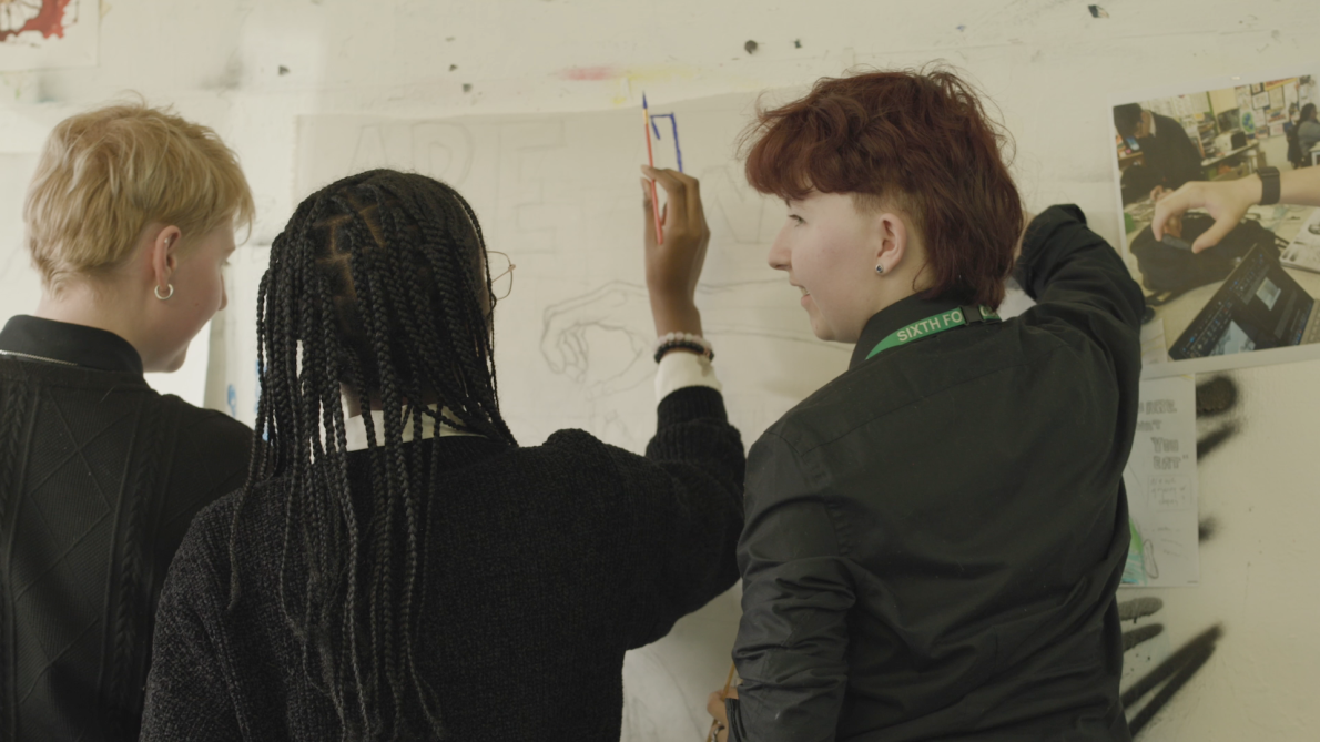 Three students collaboratively paint a large canvas on a wall.