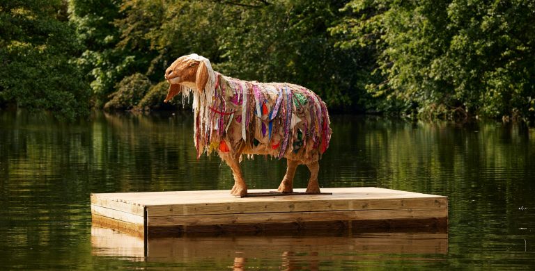A sheep sculpture, made with natural materials, seen on a floating platform in a river.