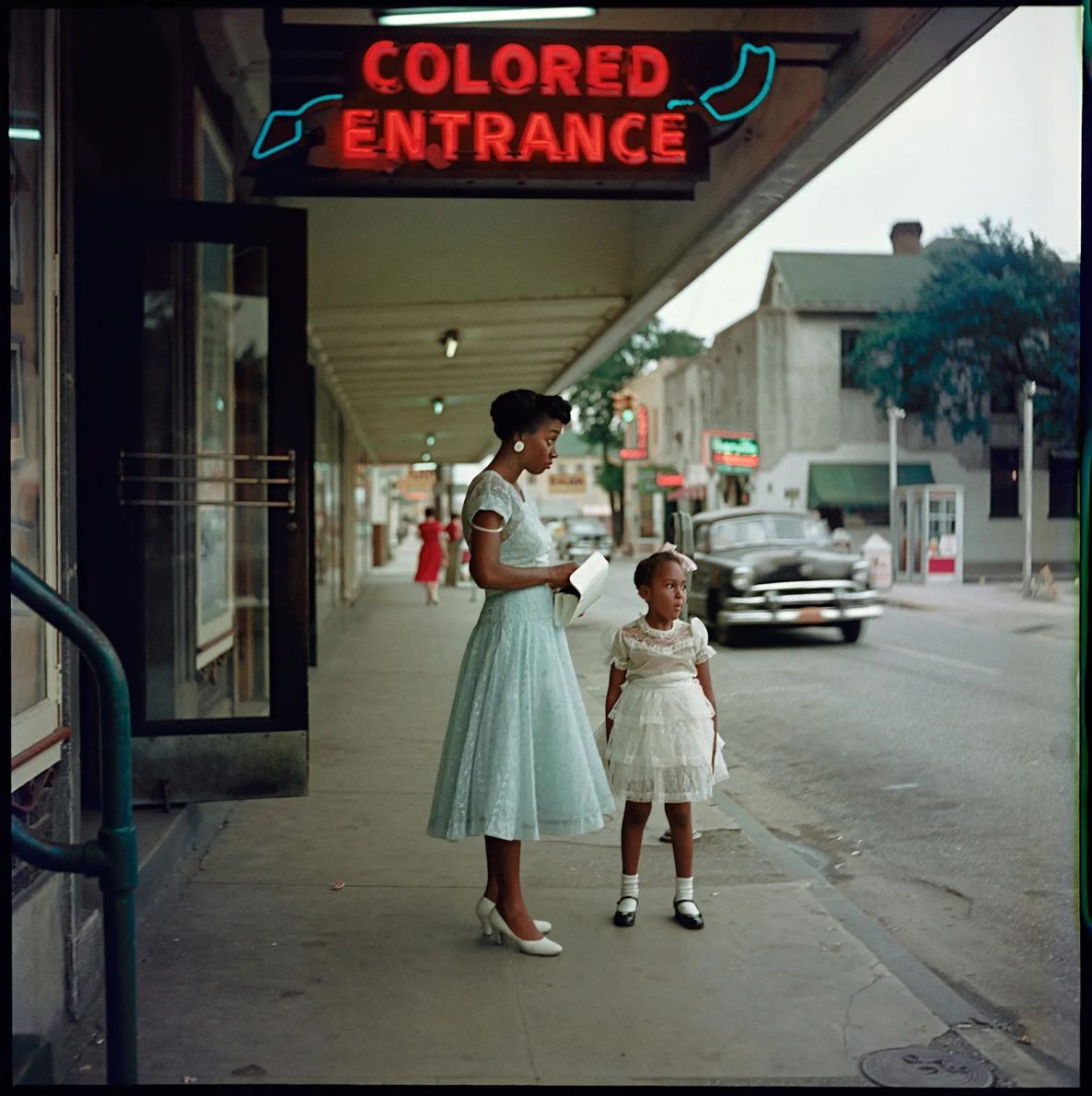 A woman in a light blue dress stands on a sidewalk next to a girl in a white dress, both looking at something off-camera. Above them, a sign reads 