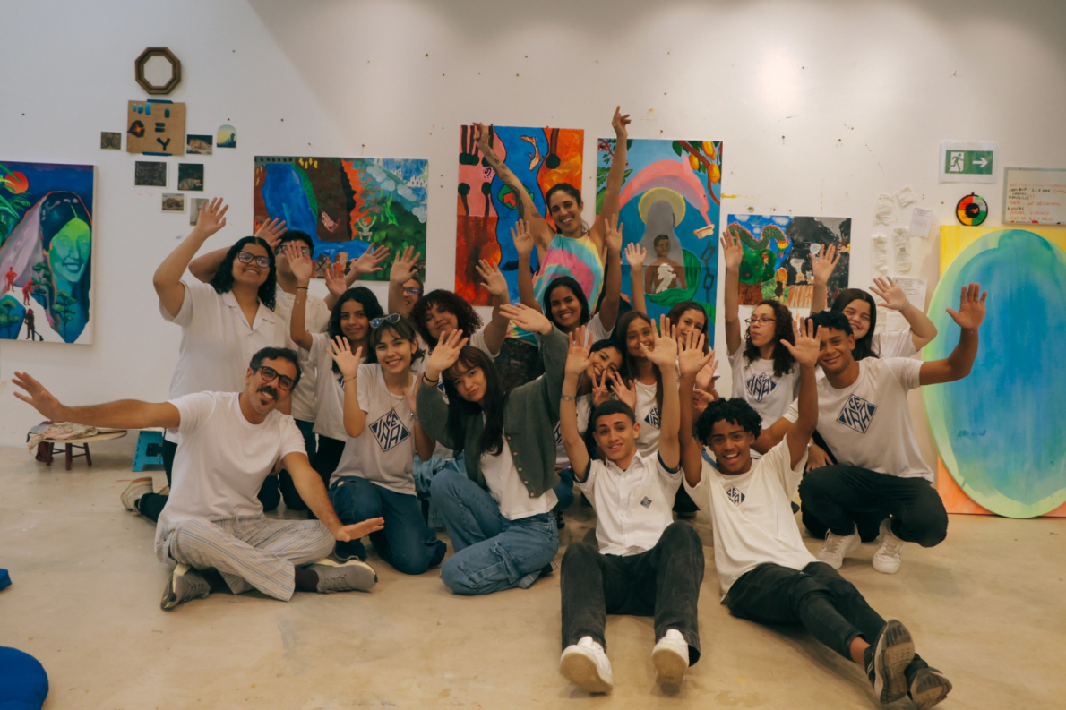 A group of smiling young people and two adults sit and kneel together in an art studio, raising their hands in celebration. Behind them, colourful abstract and figurative paintings are displayed on white walls, suggesting a collaborative art workshop or class.